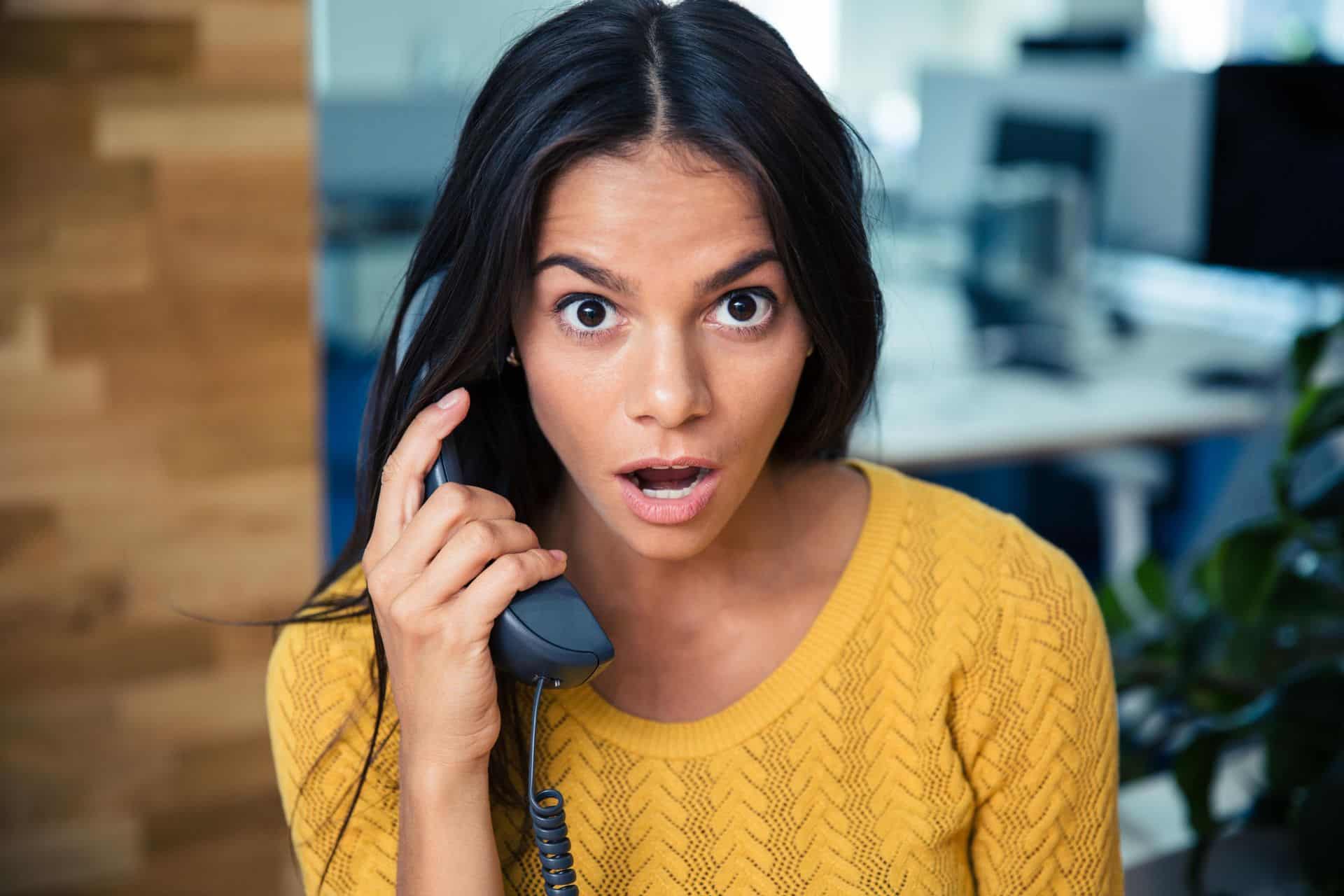 Surprise on Phone - The Sales Vault - The Sales Vault Woman in a yellow sweater speaks on a desk phone, looking surprised in an office setting.