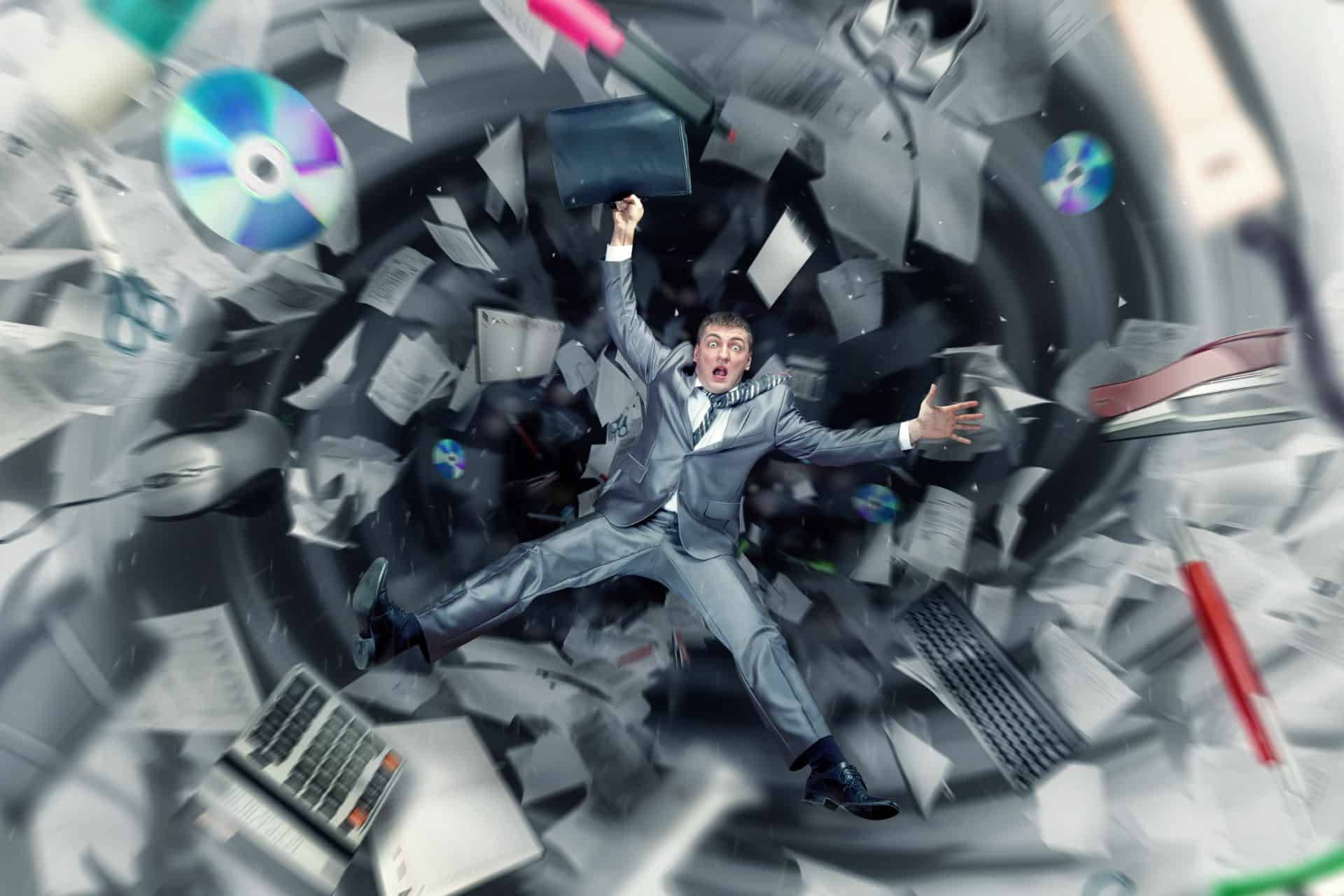 Man in a gray suit floats in a vortex of flying papers, CDs, and office gear.