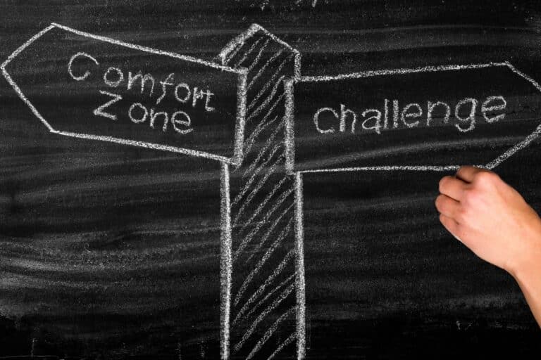 Chalk-drawn doorway from 'comfort zone' on the left to 'challenge' on the right, with a hand pushing through the barrier