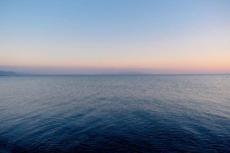 Calm ocean at dusk with a pastel gradient sky—from pale pink to deep blue—on the horizon with distant landforms barely visible.