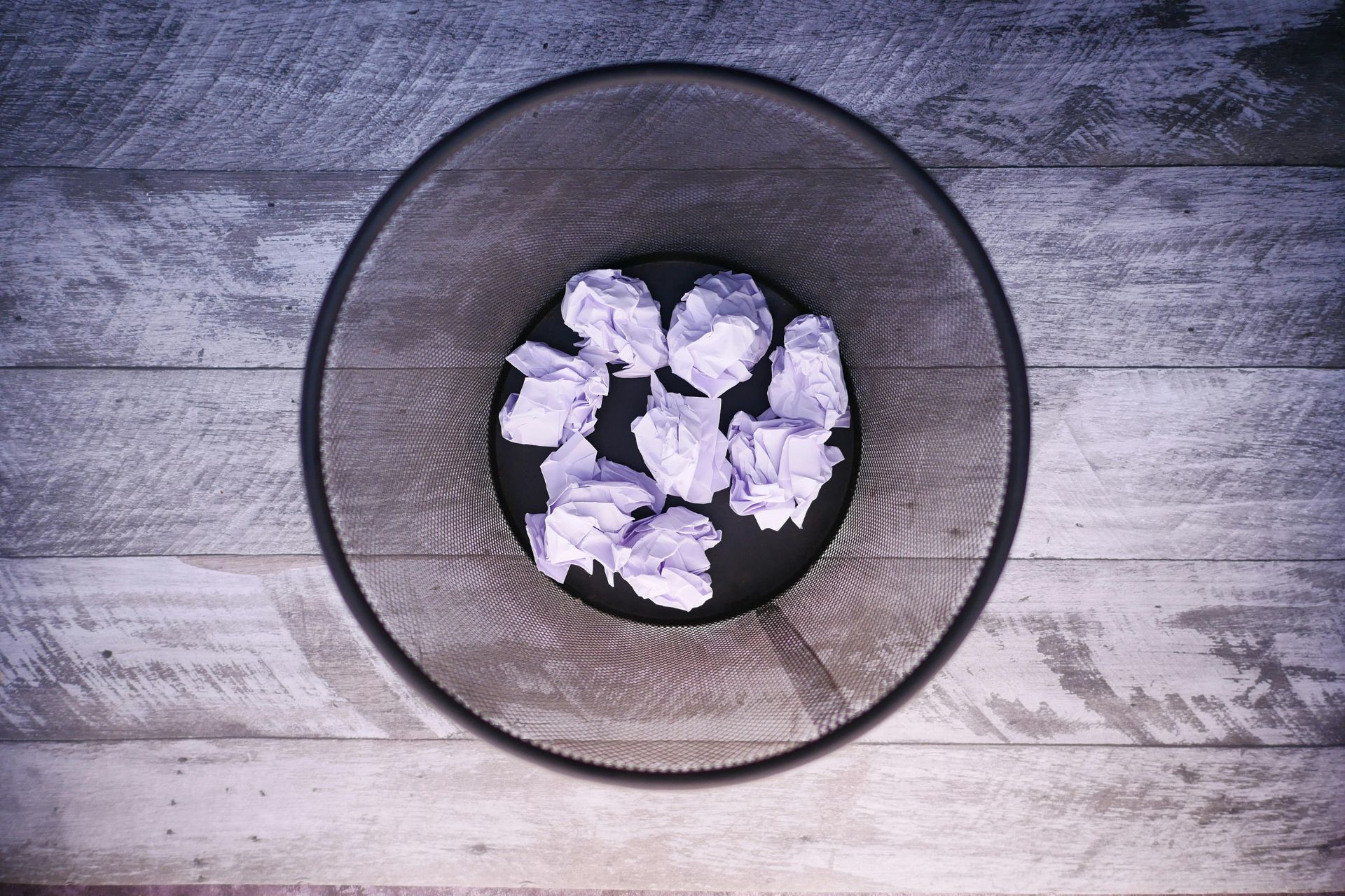 Overhead view of a black mesh wastebasket with crumpled white papers inside on a wooden floor.