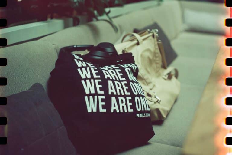 Promo Products - The Sales Vault - The Sales Vault Close-up of a black t-shirt with bold white text reading 'WE ARE ONE' on a person’s lap, next to a beige bag on a couch.
