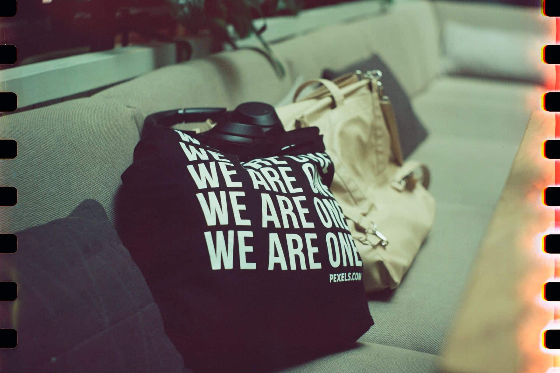 Close-up of a black t-shirt with bold white text reading 'WE ARE ONE' on a person’s lap, next to a beige bag on a couch.
