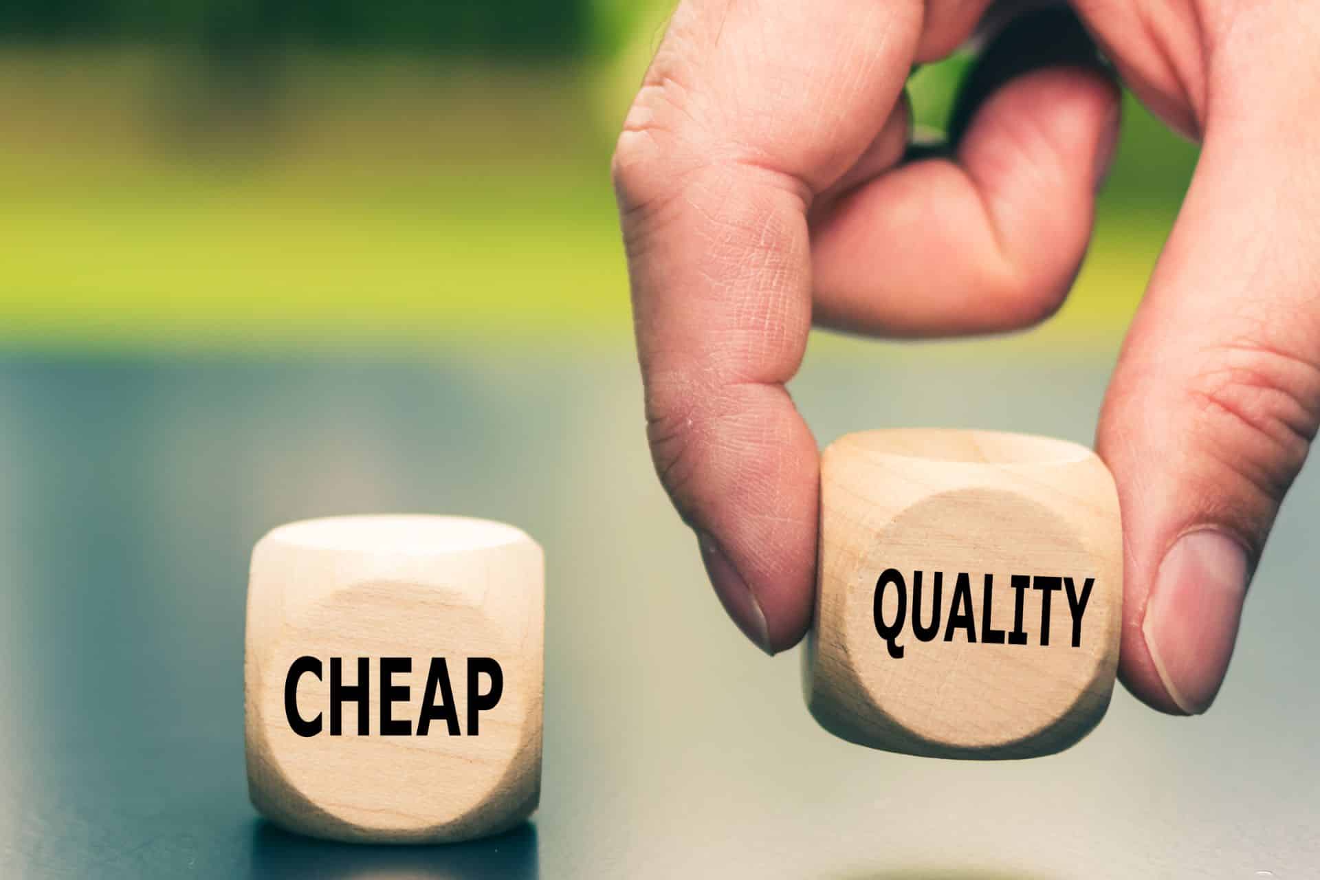 Close-up of two wooden blocks labeled CHEAP and QUALITY, held by fingers to show choosing between price and quality.