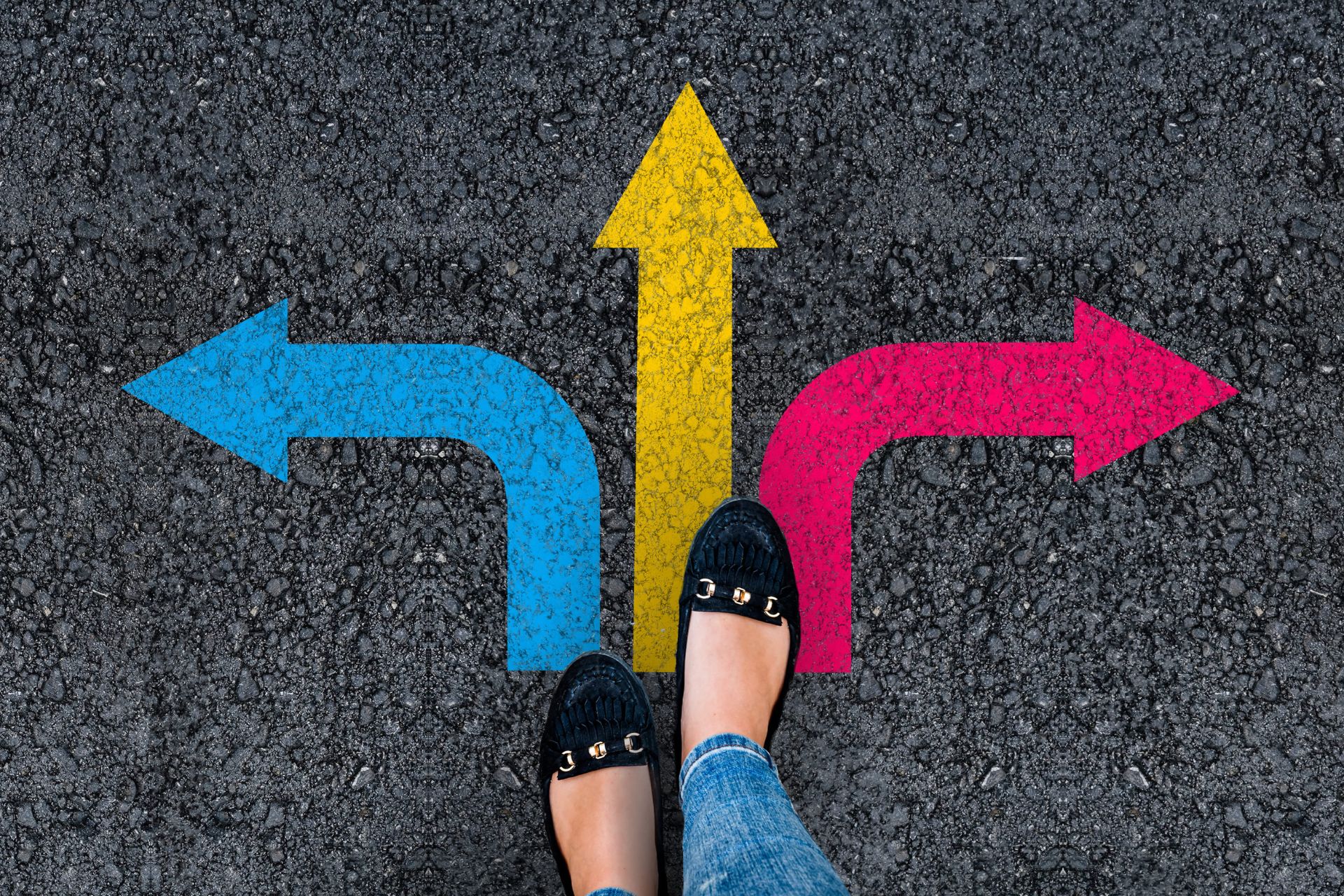 Person standing at a three-way junction with blue left arrow, yellow straight arrow, and pink right arrow painted on asphalt.