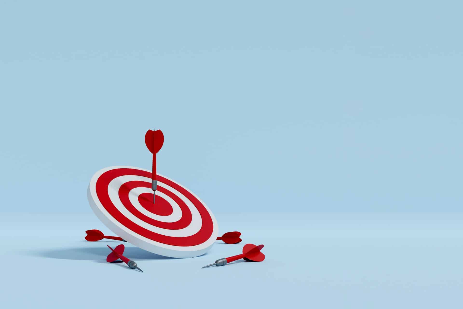 Blue background with a red-and-white bullseye target knocked over and several red darts scattered nearby on a pale blue surface.