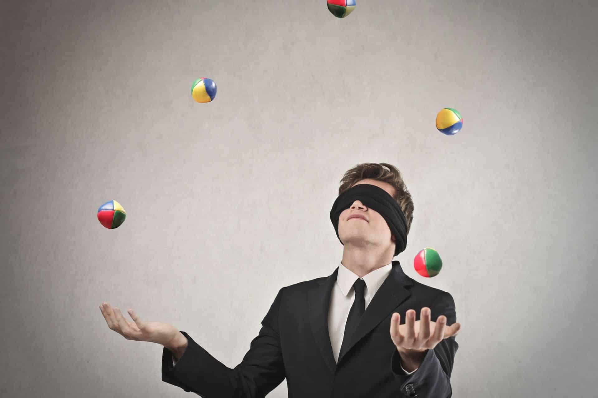 Man in a suit blindfolded while juggling colorful balls against a gray background.