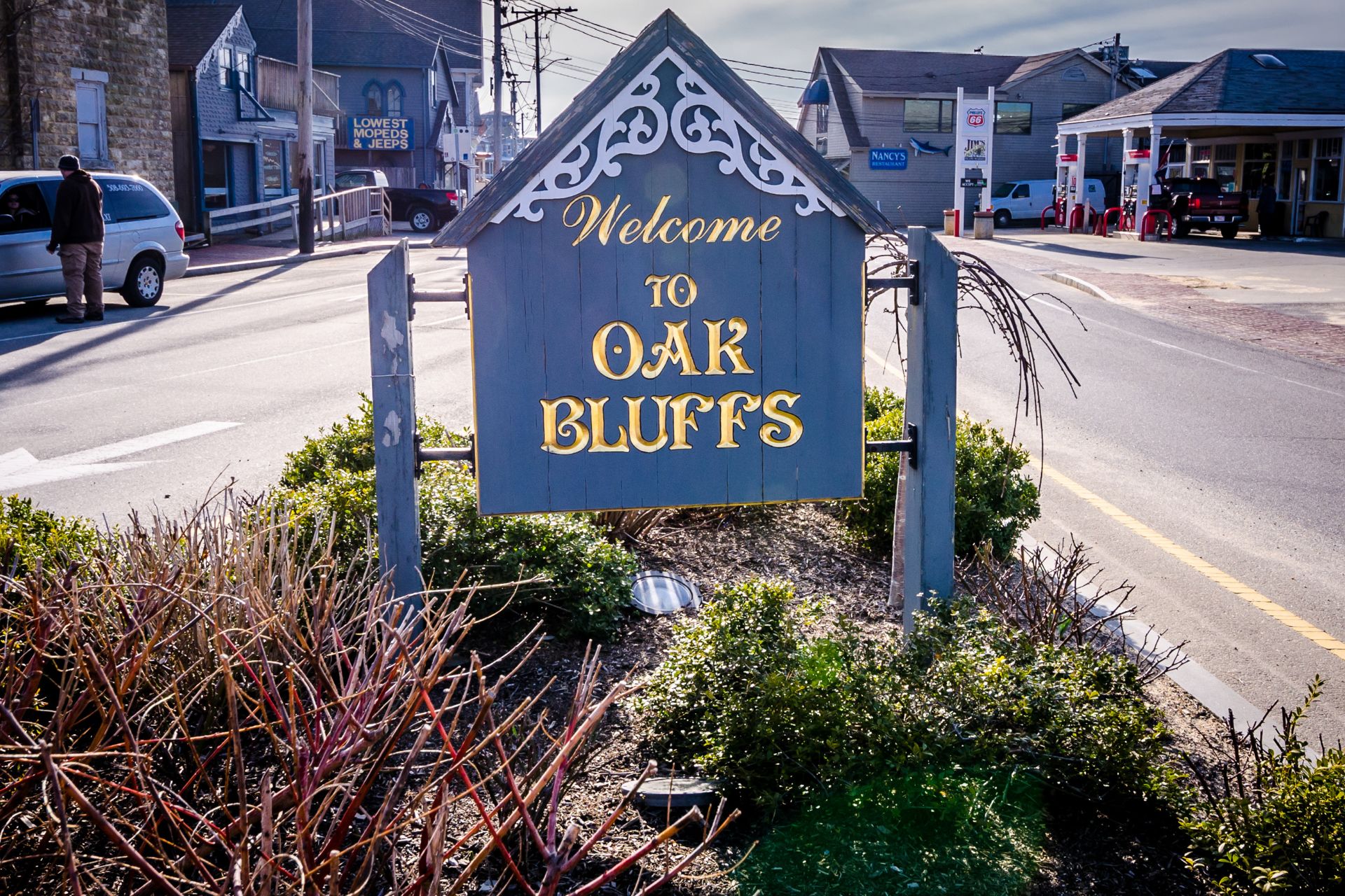 Blue wooden 'Welcome to Oak Bluffs' sign with decorative white trim and gold lettering, along a street backdrop.