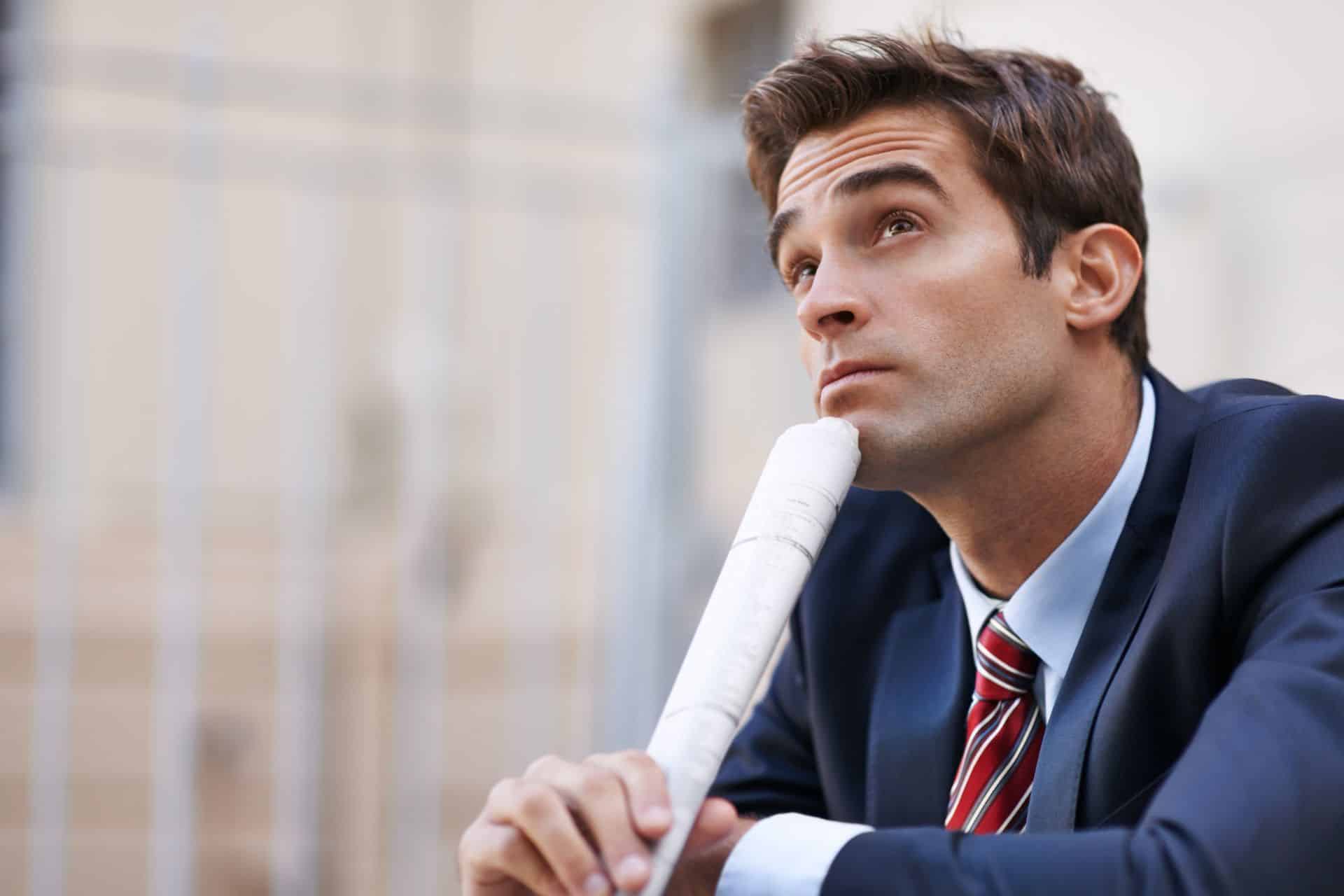 Businessman in a navy suit and striped tie looks up thoughtfully while holding a rolled blueprint.