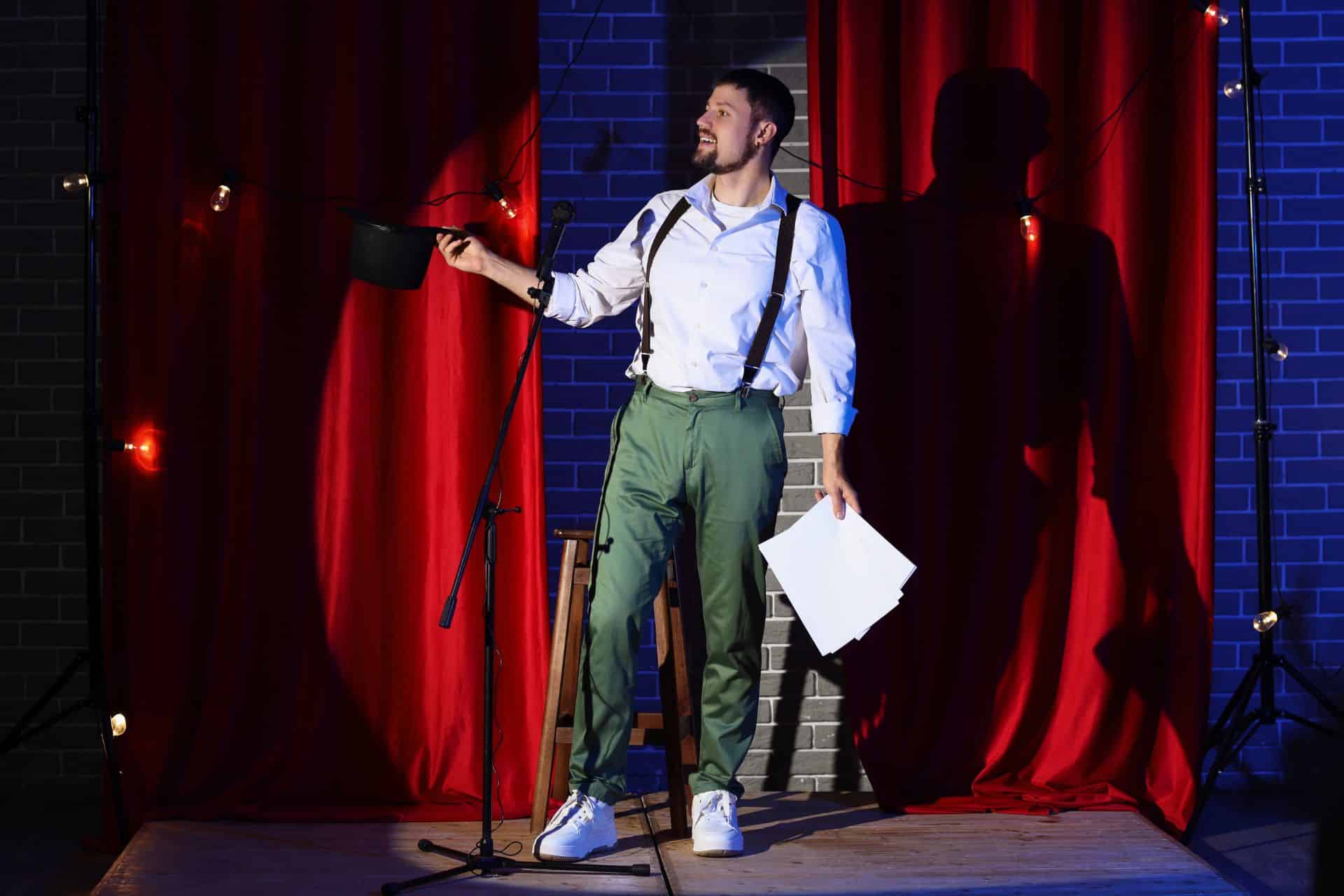 Male performer on a small stage, holding papers and a top hat, microphone in front, red curtains and brick wall behind him.