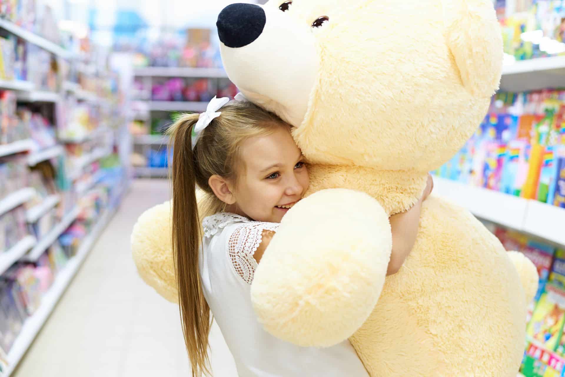 Young girl in a white dress hugs a giant beige teddy bear in a toy store aisle, smiling.