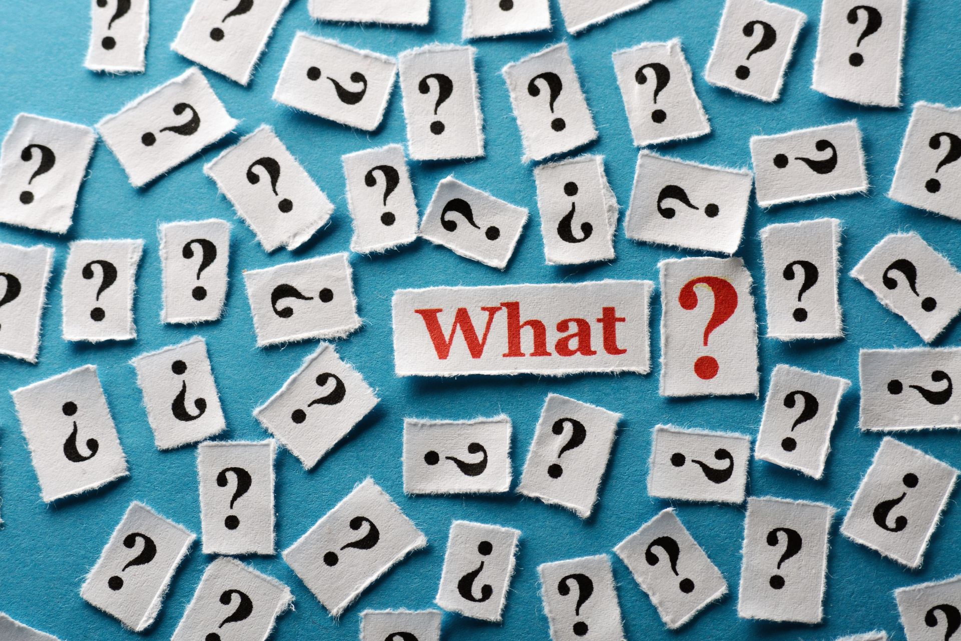 Cluster of torn white cards with black question marks scattered on a blue background, with a central card reading “What?”