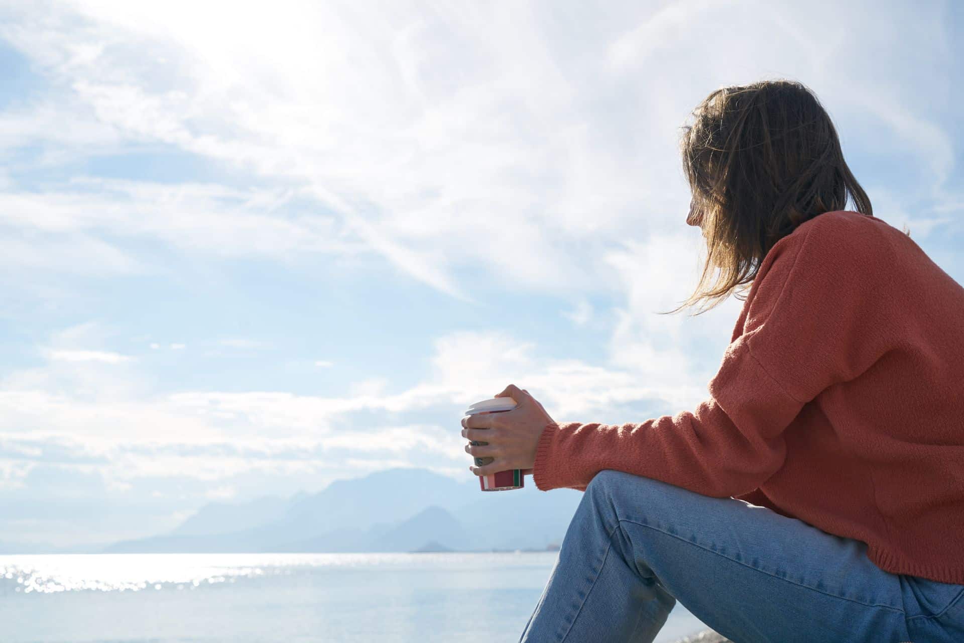 sales thoughts - The Sales Vault - The Sales Vault Person in an orange sweater sitting by the sea, holding a coffee cup, with mountains and blue sky in the background.