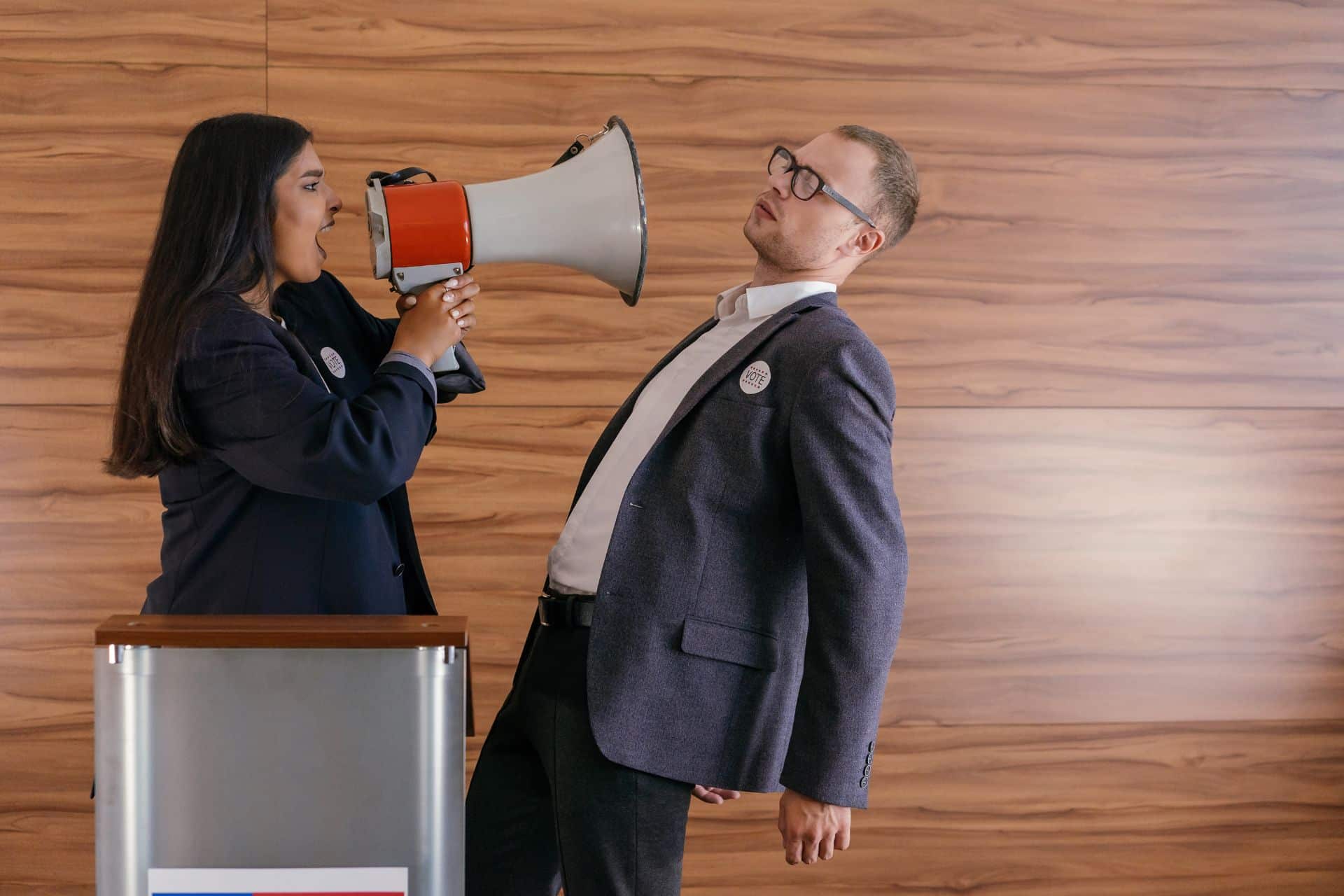 thank you sir - The Sales Vault - The Sales Vault Woman in a dark blazer yells into a red and gray megaphone aimed at a man in a suit, in a wood-paneled room (campaign/vote-themed scene).